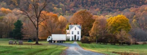 A moving truck parked in front of a charming white colonial house in the scenic hudson valley, surrounded by rolling green hills and colorful autumn foliage.